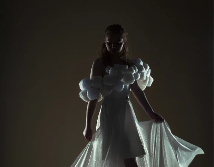 Model in a dramatic, low-lit setting wearing an off-the-shoulder white gown with textured puff sleeves and a flowing sheer train, softly illuminated from behind. Model in a dramatic, low-lit setting wearing an off-the-shoulder white gown with textured puff sleeves and a flowing sheer train, softly illuminated from behind.